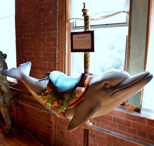 A colorful dolphin carousel figure with a saddle, displayed indoors against a brick wall and a window.