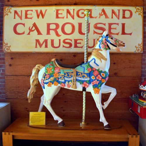 A colorful carousel horse displayed at the New England Carousel Museum, with a decorative sign in the background.