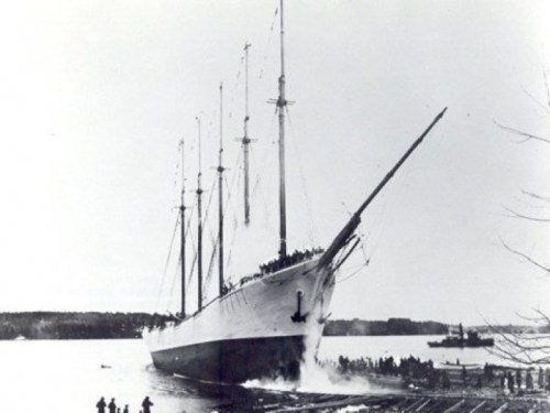 Historic black-and-white photo of a large sailing ship launching into water, with spectators watching from the shore.
