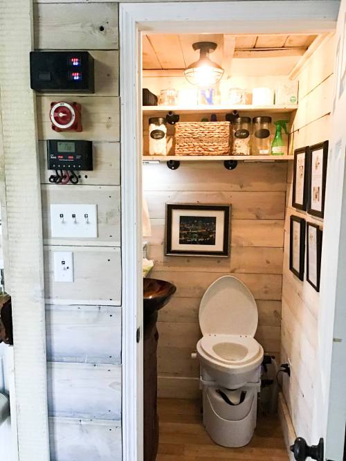 A small bathroom with wooden walls, featuring a toilet, shelves with jars, and framed pictures.