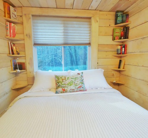 Cozy bedroom with a wooden interior, a large bed, and bookshelves filled with books beside a window.