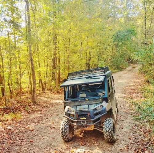 A black off-road vehicle parked on a dirt trail surrounded by trees with autumn foliage.