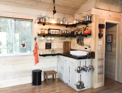 Cozy kitchen with wooden walls, open shelves, a sink, and pots hanging, featuring natural light from a large window.