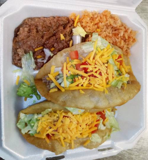 Two crispy tacos filled with lettuce, tomatoes, and cheese, served with refried beans and rice in a takeout container.