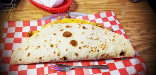 A large, folded tortilla filled with yellow cheese and meat, served on a red and white checkered plate with a fork.