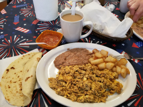 A plate with scrambled eggs, refried beans, diced potatoes, tortillas, and a cup of coffee on a festive tablecloth.