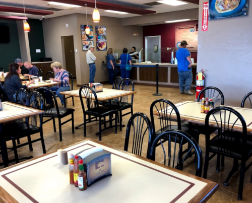 A casual restaurant interior with empty tables, patrons at the counter, and condiments on the tables.