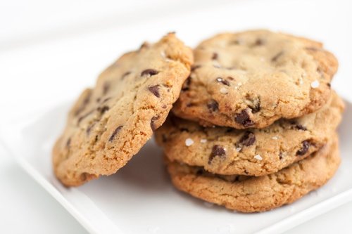 A stack of chocolate chip cookies on a white plate, with a sprinkle of sea salt on top.