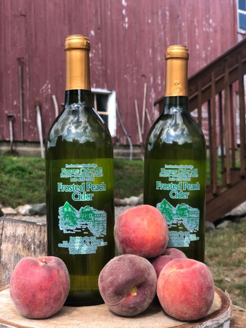Two bottles of Frosted Peach Cider beside fresh peaches, set against a rustic red barn and wooden steps.
