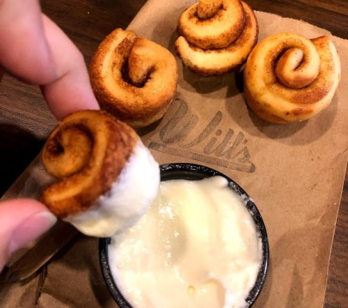 A hand holding a cinnamon roll with three more rolls and a small cup of icing on a brown paper surface.