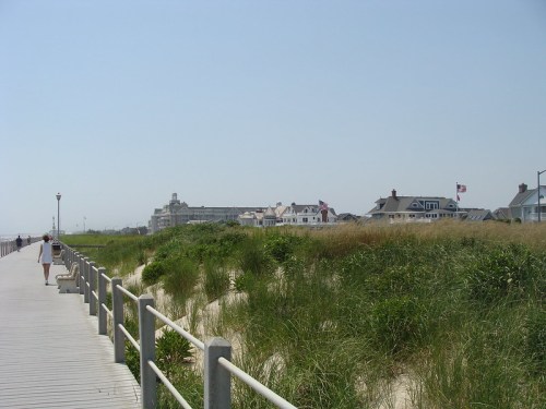A wooden boardwalk lined with tall grass, leading to beach houses under a clear blue sky.
