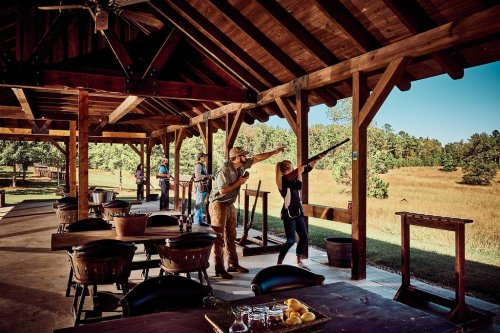 A rustic outdoor shooting range with people practicing shooting under a wooden pavilion surrounded by nature.