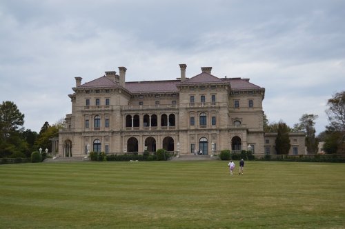 A grand mansion with ornate architecture, set against a cloudy sky, with two people walking on a grassy lawn.