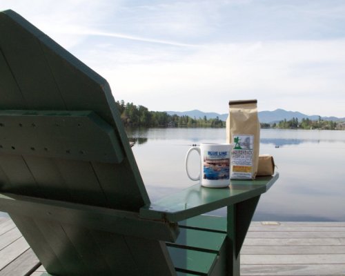 A green Adirondack chair by a calm lake, with a coffee mug and a bag of coffee on the armrest, mountains in the background.