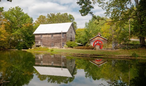 A rustic wooden mill and a red waterwheel shed reflect in a calm pond, surrounded by trees and a cloudy sky.