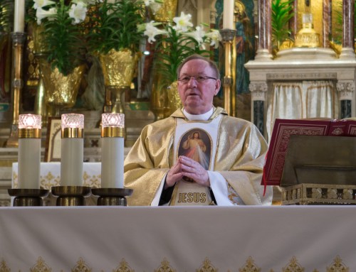 A priest in a gold vestment stands at an altar, holding a religious image, with candles and floral decorations nearby.