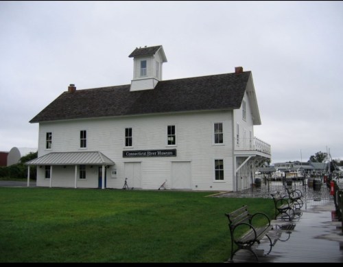 White building with a cupola, labeled "Connecticut River Museum," near a marina and grassy area on a cloudy day.