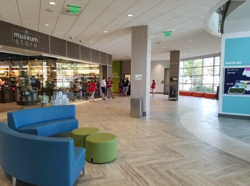 Interior of a museum lobby featuring a store, seating area, and visitors looking at a map. Bright and spacious design.