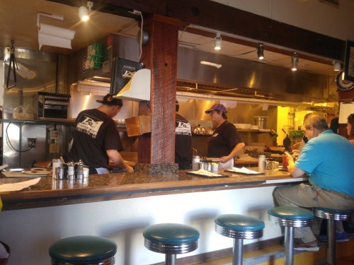 A busy diner kitchen with staff cooking, and a customer seated at the counter, enjoying the atmosphere.