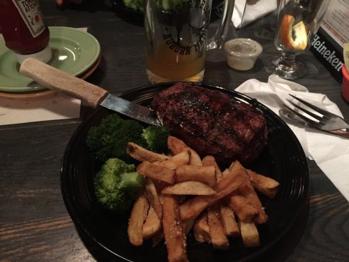 A plate with grilled steak, broccoli, and French fries, accompanied by a beer and condiments on a wooden table.