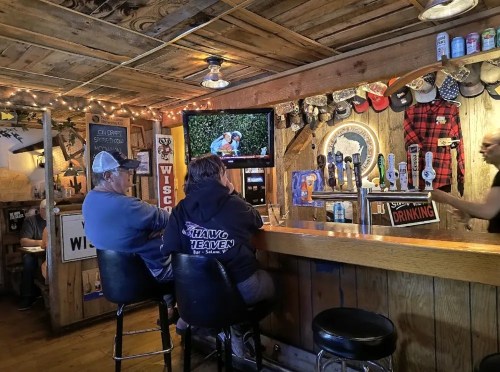 Two patrons sit at a rustic bar with a TV showing a sports event, surrounded by wooden decor and beer taps.