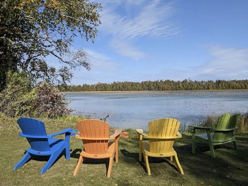 Four colorful Adirondack chairs by a serene lake, surrounded by trees and a clear blue sky.
