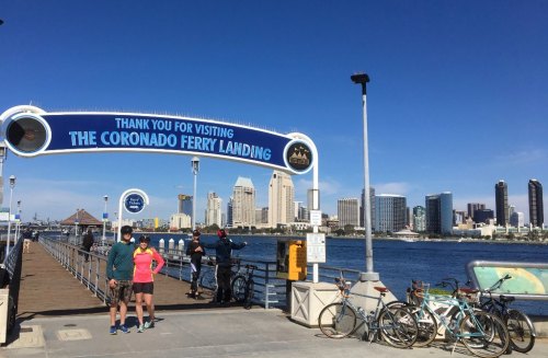Two people stand on a pier at Coronado Ferry Landing, with city skyline and bicycles in the background under a blue sky.
