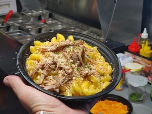 A hand holding a bowl of macaroni and cheese topped with pulled meat and breadcrumbs, with condiments in the background.