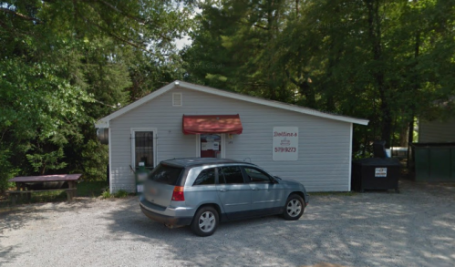 A small gray building surrounded by trees, with a parked car in front and a dumpster nearby.