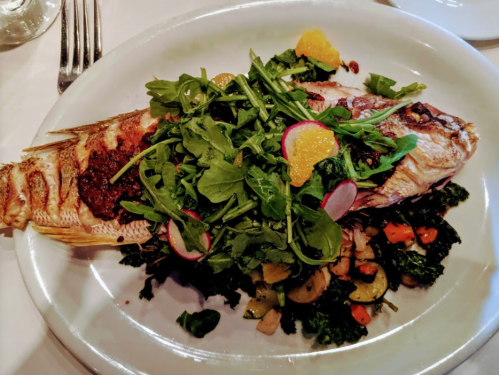 A whole grilled fish topped with arugula, radishes, and orange slices, served on a bed of sautéed vegetables.