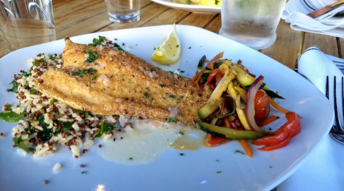 A plate of grilled fish with lemon, served with quinoa and a colorful vegetable medley on a wooden table.