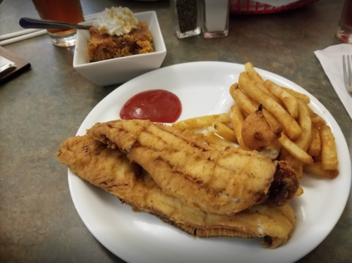 A plate with two fried fish fillets, French fries, a side of ketchup, and a dessert in a bowl.