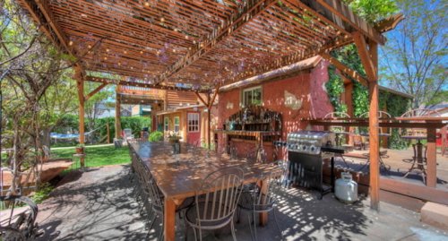 Outdoor dining area with a wooden table, chairs, and a grill, shaded by a pergola surrounded by greenery.