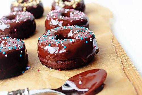 A close-up of chocolate-glazed donuts topped with colorful sprinkles on a parchment-lined wooden surface.