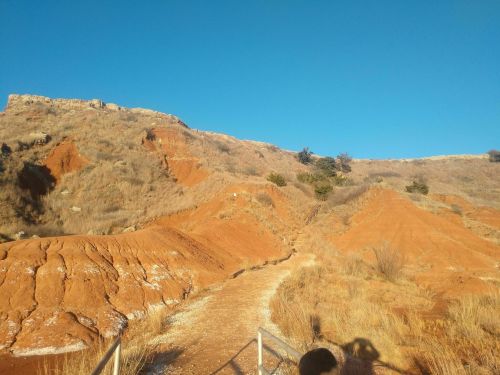 A winding dirt path leads through reddish-brown hills under a clear blue sky. Sparse vegetation is visible.