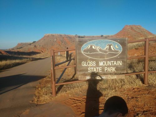 Sign for Gloss Mountain State Park with red rock formations in the background and a clear blue sky.