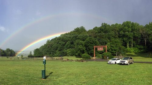 A double rainbow arcs over a green landscape with two parked cars and a wooden entrance sign in the background.