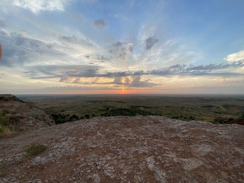 A scenic view of a sunset over a vast landscape, with clouds scattered across the sky and rolling hills in the foreground.