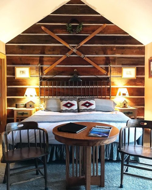 Cozy bedroom with a wooden accent wall, a bed with decorative pillows, and two chairs around a small table.