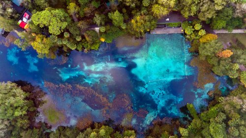 Aerial view of a vibrant blue and green river surrounded by lush trees and a pathway along the bank.