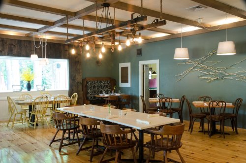 Cozy restaurant interior with wooden tables, chairs, and warm lighting, featuring a green wall and decorative branches.
