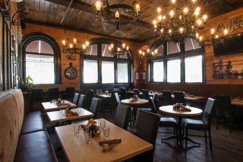 Cozy restaurant interior with wooden walls, chandeliers, and tables set for dining, featuring large windows and soft lighting.