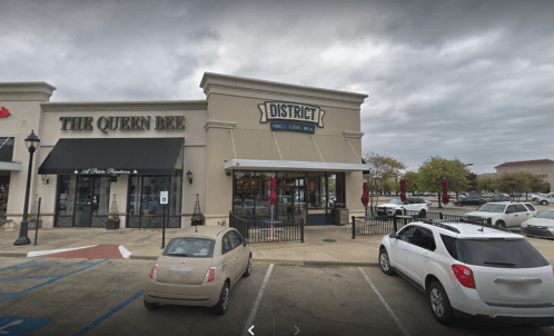 Exterior view of a shopping area featuring "District" restaurant and "The Queen Bee" boutique, with parked cars in front.