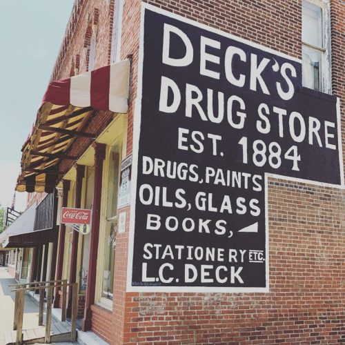 A vintage brick building with a large sign reading "Deck's Drug Store, Est. 1884" and a Coca-Cola sign nearby.