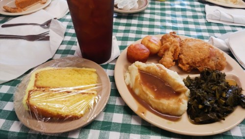 A plate with fried chicken, mashed potatoes, collard greens, cornbread, and a glass of iced tea on a checkered tablecloth.