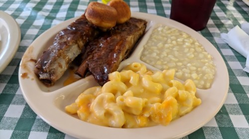 A plate of barbecue ribs with cornbread, macaroni and cheese, and corn pudding on a checkered tablecloth.