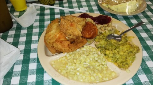 A plate of fried chicken, cornbread, corn, green beans, stuffing, and cranberry sauce on a checkered tablecloth.