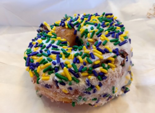 A glazed donut topped with colorful yellow, green, and purple sprinkles on a white background.