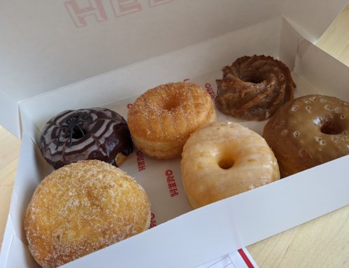 A box of six assorted donuts, including chocolate, glazed, and cinnamon varieties, on a light wooden surface.