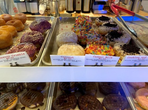 A display of colorful donuts with various toppings, labeled with prices, in a bright bakery setting.